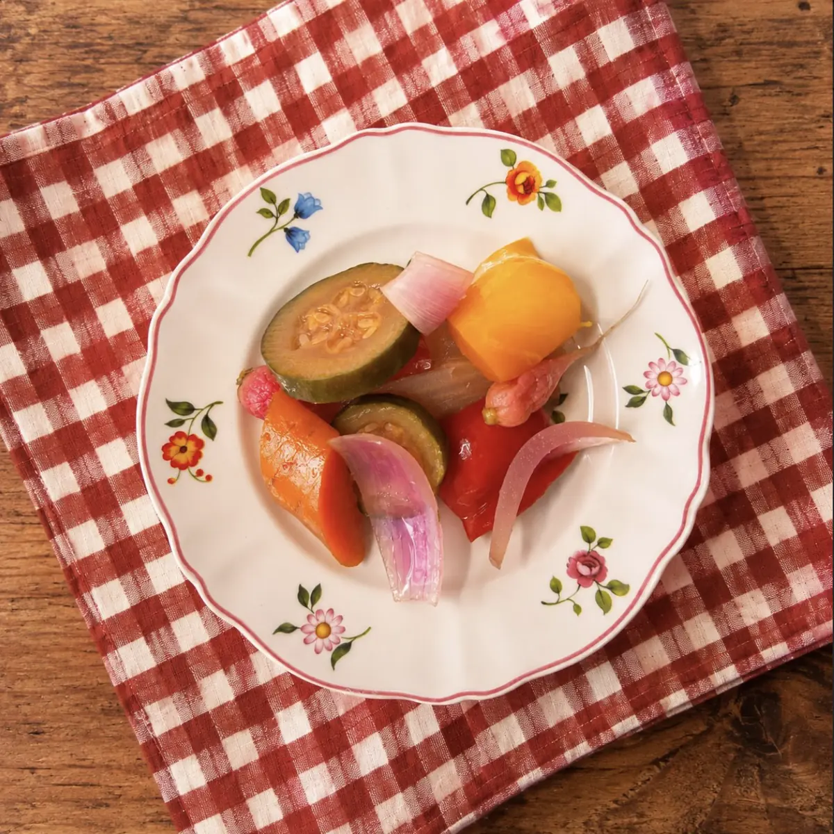 Giardiniera di verdure — zucchine, carote, peperone, cipollotto — su un piatto a fiori, sopra un tovagliolo a quadretti rossi.