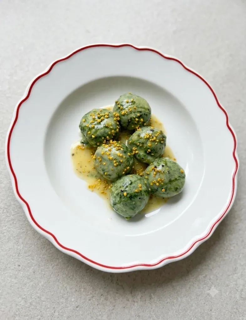 Gnudi verdi di ricotta e spinaci nel burro, cosparsi di polline, su un piatto bianco dal bordo rosso.